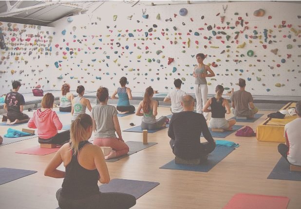 Clase de yoga para escaladores frente a un muro de bloque, con el grupo sentado sobre esterillas